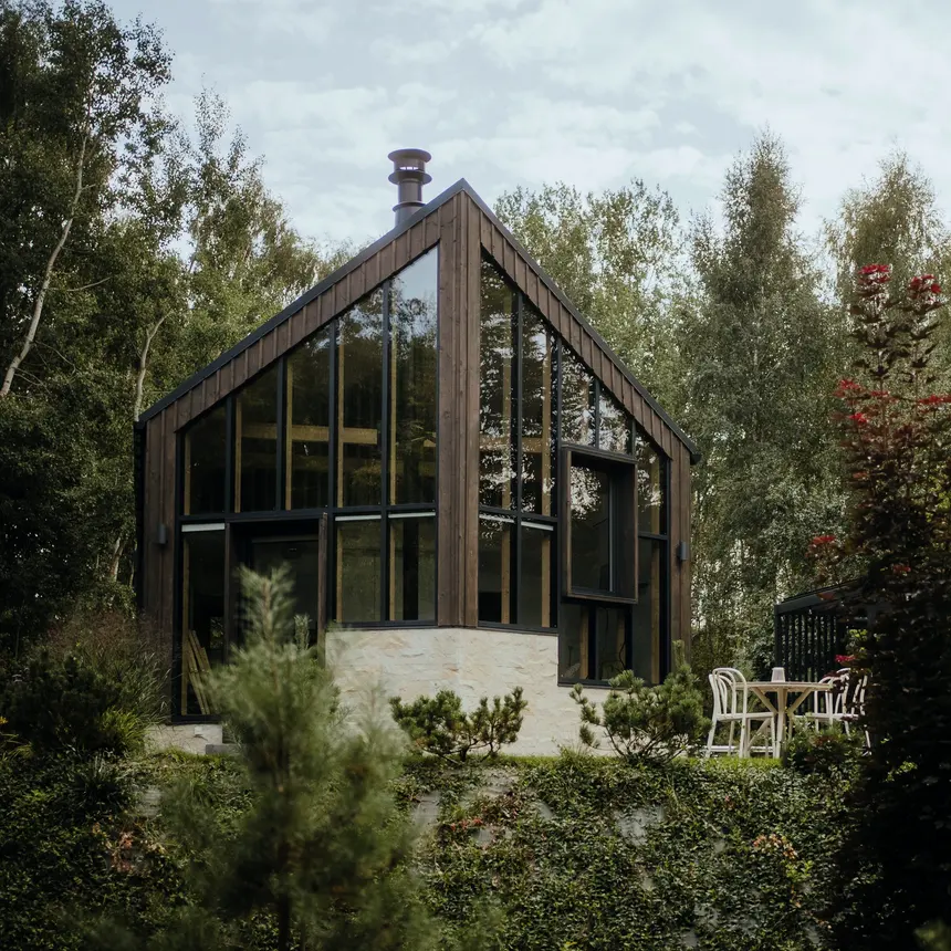 A wooden cabin at dusk with a string of lights and warmly lit windows among greenery